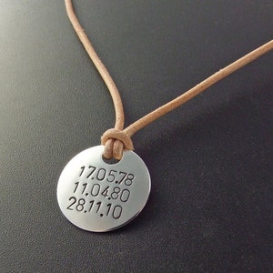 May include: A silver pendant necklace with a brown leather cord. The pendant is round and has the dates 17.05.78, 11.04.80, and 28.11.10 engraved on it.