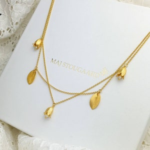 Könnte beinhalten: Eine goldfarbene Halskette mit mehrschichtigem Design. Die Halskette hat kleine Anhänger in Form von Blumen und Blättern. Die Halskette wird auf einer weißen Schachtel mit dem Text "MAJ STOUGAARD" präsentiert.