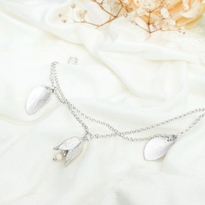 Könnte beinhalten: Ein zartes silbernes Armband mit einem Blumenanhänger und zwei Blattanhängern. Das Armband besteht aus zwei Ketten und hat einen Perlenakzent am Blumenanhänger.