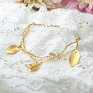 Könnte beinhalten: Ein zartes goldenes Kettenarmband mit einem kleinen Blumenanhänger und zwei Blattanhängern. Das Armband ist mehrlagig und hat einen zarten, femininen Look.