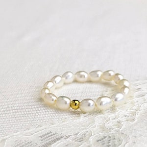 Könnte beinhalten: Ein weißes Perlenarmband mit einem einzigen goldenen Perlenakzent. Das Armband liegt auf einem weißen Spitzenhintergrund.