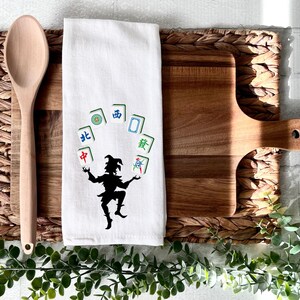 May include: White kitchen towel with a black silhouette of a jester juggling mahjong tiles. The tiles are green, blue, and red with Chinese characters.
