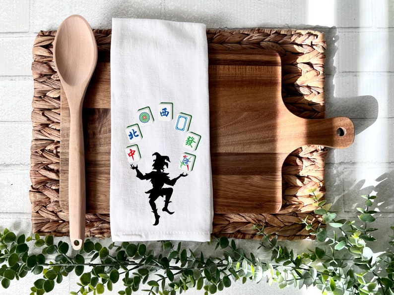 May include: White kitchen towel with a black silhouette of a jester juggling mahjong tiles. The tiles are green, blue, and red with Chinese characters.