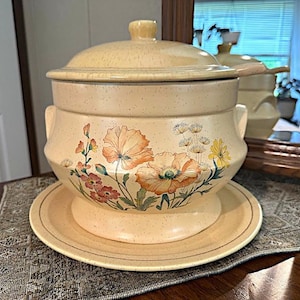 May include: Cream-colored ceramic soup tureen with lid, handles, and matching plate. The tureen and plate feature a floral design in shades of orange, pink, and yellow. The tureen is speckled and sits on a matching plate, creating a cohesive set.