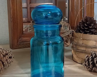 May include: A clear, turquoise glass apothecary jar with a matching round lid. The jar has a wide base and a narrower neck, with a lid that fits snugly on top. The jar is displayed on a neutral surface, with a wooden mirror and decorative items in the background.
