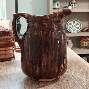May include: A brown ceramic pitcher with a handle and spout. The pitcher has a glossy finish with dark and light brown tones. The surface features a textured pattern. The pitcher is likely used for serving beverages.