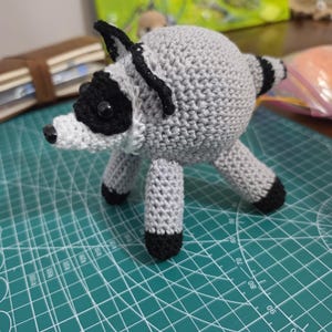 May include: A round, crocheted raccoon plush toy featuring a light grey body, a white muzzle, black eye patches, black ears, and a striped tail. The raccoon has four legs with black tips and small, dark, round eyes, crafted with visible yarn stitches.