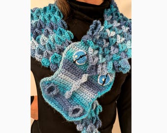 May include: A blue and teal crocheted dragon scarf. The scarf features a dragon head with blue eyes and a snout. The scarf is made of various shades of blue yarn, creating a textured, scaly appearance. The dragon's head is the focal point.