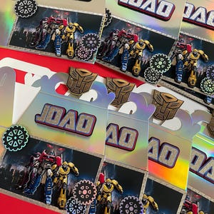 May include: Party favor boxes with a Transformers theme. Each box features the name "JOAO" in silver letters, along with images of Transformers characters, cogwheel accents, and the Autobot symbol. The boxes have a silver and red color scheme.