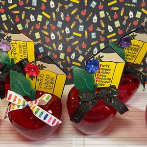 Puede incluir: Contenedores rojos en forma de manzana con detalles de hojas verdes y lazos decorativos. Cada manzana tiene una etiqueta en forma de lápiz que dice "Gracias por ser mi profesor". El fondo presenta patrones con temática escolar.