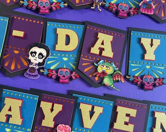 May include: A colorful banner with the words "Happy Day" and "Even" in gold glitter letters. The banner is decorated with purple, blue, and orange paper cutouts and features characters from the movie Coco.