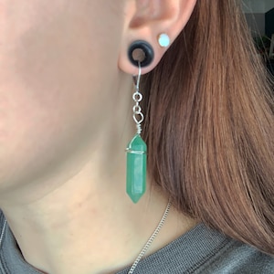 Könnte beinhalten: Ein silberner Kettenohrring mit einem grünen Kristallanhänger. Der Kristall ist facettiert und hat eine spitze Spitze. Der Ohrring hängt am Ohrläppchen einer Person.