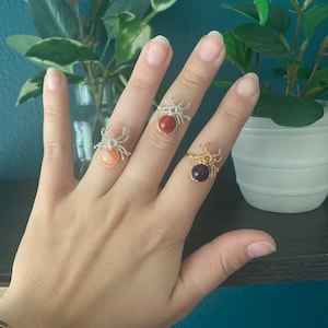 以下が含まれることがあります： クモのデザインとカラフルな宝石が付いた銀と金の指輪3つ。指輪は人の手につけられています。