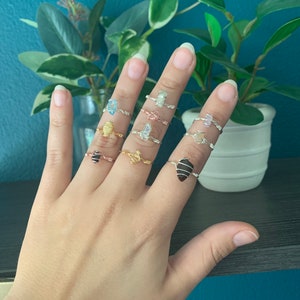 May include: A hand wearing eight different wire-wrapped rings with various colored stones. The rings are made with silver and gold wire.