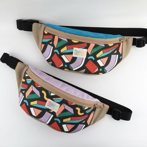 Könnte beinhalten: Zwei Bauchtaschen mit einem geometrischen Muster in Rot, Orange, Türkis, Lila und Gelb auf schwarzem Hintergrund. Die Bauchtaschen haben eine beige Basis, einen schwarzen verstellbaren Riemen und einen Reißverschluss. Die Marke "HOPPUS" ist auf einem Etikett.