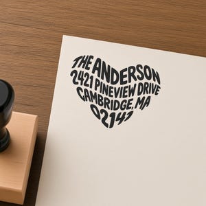 May include: A wooden rubber stamp with a black handle and a square base, shown next to a cream-colored paper featuring a black ink imprint of the text THE ANDERSON 2421 PINEVIEW DRIVE CAMBRIDGE, MA 02147 arranged in the shape of a heart.