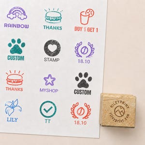 May include: A wooden rubber stamp with a circular logo reading RILEYPRINT and rileyprint.com, placed next to a white sheet featuring twelve colourful ink stamp impressions including icons of a rainbow, burger, drink, paw, heart, star, lily, and text.
