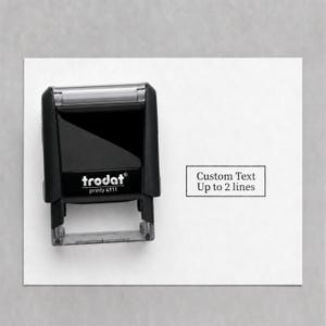 May include: A black Trodat Printy 4911 self-inking stamp with a rectangular body and a clear base. Next to the stamp, a white rectangular area displays the black printed text Custom Text Up to 2 lines inside a thin black rectangular border.