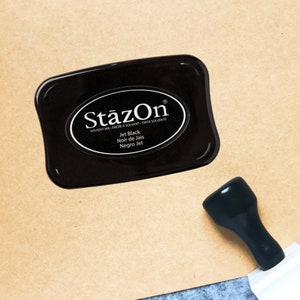May include: A black StāzOn ink pad with the text "StāzOn" and "Jet Black" in white lettering. A black stamp handle is visible to the right of the ink pad. The ink pad is on a tan surface.