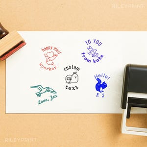 May include: A collection of custom rubber stamps with various designs and text, including a heart with mail, a bird with a letter, a snail with an envelope, a squirrel, and a seagull. The stamps are in red, blue, and green ink.