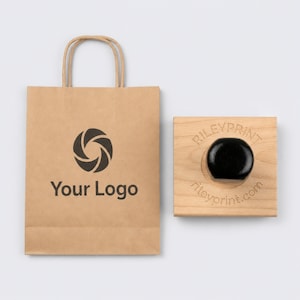 May include: A brown paper shopping bag with handles featuring a black circular aperture logo and the text Your Logo, alongside a square wooden rubber stamp with a black handle and the text RILEYPRINT rileyprint.com engraved on the wood surface.