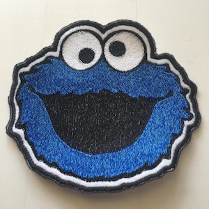 May include: Embroidered patch featuring the Cookie Monster from Sesame Street. The patch has a blue, furry design with a large, open black mouth and two large white eyes with black pupils. The patch is outlined in black and white.