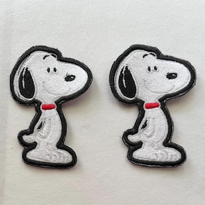 May include: Two embroidered patches featuring Snoopy from the Peanuts comic strip. Each patch depicts Snoopy in a standing pose, with a white body, black ears, and a red collar. The patches have a black border.