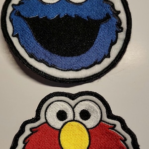 May include: Two embroidered patches featuring Sesame Street characters. One patch depicts Cookie Monster with a blue face, black mouth, and white eyes. The other patch shows Elmo with a red face, black mouth, and yellow nose.