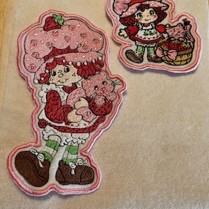 Puede incluir: Dos parches bordados que muestran un personaje de pelo rojo con un gran sombrero rosa con estampado de fresas, un vestido rojo y calcetines a rayas verdes. Un parche muestra al personaje sosteniendo un gato rosa y el otro una cesta de fresas.