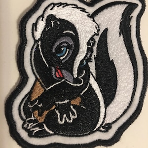 May include: Embroidered patch featuring a cartoon skunk with black fur, a white stripe, and a fluffy white tail. The skunk has blue eyes, a red tongue, and is surrounded by a white border with black stitching. The patch is likely for clothing or accessories.