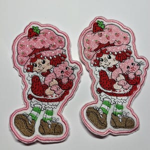 May include: Two embroidered patches featuring a character with red hair wearing a pink strawberry-patterned hat, a red sweater, and green and white striped socks, while holding a small pink cat. The patches have a pink border and white backing.