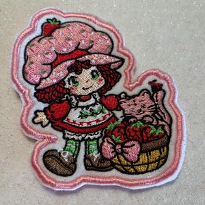 May include: An embroidered patch featuring a character with red hair wearing a pink strawberry-patterned hat, a red dress with a white apron, and green striped stockings. Beside the character is a pink cat sitting next to a basket of red strawberries.