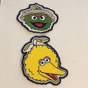 May include: Two embroidered patches featuring Sesame Street characters. One patch depicts Oscar the Grouch in a tan hat, with green fur and a red mouth. The other patch shows Big Bird, a yellow bird with a long beak and large eyes.