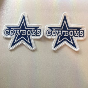 Może przedstawiać: Dwie haftowane naszywki z niebieskim wzorem gwiazdy i napisem "COWBOYS" w białych literach. Naszywki mają białą obwódkę. Tło jest jasne. Te naszywki są idealne do dodania do odzieży lub akcesoriów.