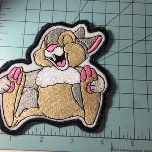 May include: Embroidered patch featuring a cartoon rabbit with a joyful expression. The patch has a black border and includes shades of tan, gray, pink, and white. The rabbit is lying down with its paws up. The patch is on a green cutting mat.
