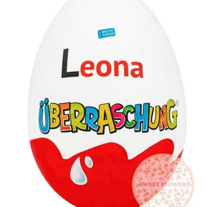 Könnte beinhalten: Ein weißes Plastikei mit einem rot-weißen Schokoladen-Design. Das Ei hat die Worte "Überraschung" und "Leona" aufgedruckt. Das Ei ist eine beliebte deutsche Schokoladen-Spezialität.