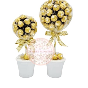 Könnte beinhalten: Zwei dekorative Arrangements aus goldverpackten Schokoladenbonbons, die kleinen Bäumen ähneln. Jedes Arrangement steht in einem weißen Topf und ist mit einem goldenen Band gebunden. Die Süßigkeiten sind zusammengefasst und bilden eine ansprechende Präsentation.