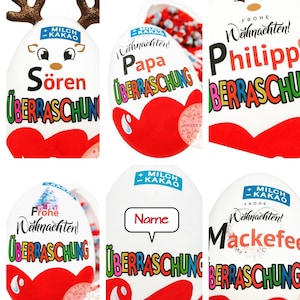 Könnte beinhalten: Sechs rote und weiße Schokoladeneier mit einer Überraschung im Inneren. Jedes Ei hat ein anderes Design mit einem Weihnachtsmotiv. Die Eier haben die Worte "Frohe Weihnachten!" und "Überraschung" darauf. Die Eier sind mit Rentiergeweihen, einem Weihnachtsbaum und einem Namen verziert.
