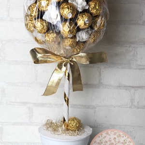 Könnte beinhalten: Eine dekorative Anordnung, die einem Baum ähnelt, mit goldverpackten Pralinen und weißen floralen Akzenten. Die Pralinen sind in einer transparenten Zellophanverpackung gruppiert, die mit einem goldenen Band gebunden ist. Die Basis ist ein weißer Topf mit goldenem Akzent.