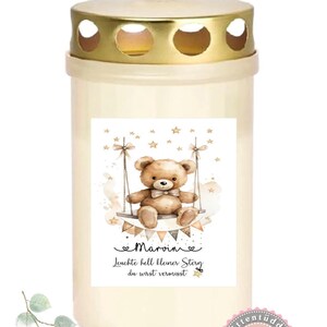 Könnte beinhalten: Weiße Gedenkkerze mit goldenem Deckel. Die Kerze hat einen Aufkleber mit einem Teddybären auf einer Schaukel, Sternen und dem Text "Marvin Leuchte hell kleiner Stern du wirst vermisst".