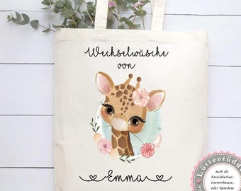 May include: A white canvas tote bag with a cute giraffe illustration and the text "Wechselwäsche von Emma" printed on it. The bag has a floral wreath around the giraffe and a pink flower on its head. The bag also has a small logo in the bottom right corner that says "Lüttentuddel" with a website address.