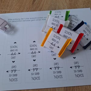 Könnte beinhalten: Ein Blatt Papier mit aufgedrucktem Text und mehreren kleinen, verpackten Süßigkeiten. Der Text auf dem Papier lautet "SCHON, dass es dich Gibt!" und "Bitte entlang der Linie ausschneiden". Die Süßigkeiten sind in weißes Papier mit farbigen Bändern verpackt.