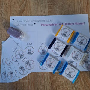 Può includere: Un assortimento di articoli a tema pasquale, tra cui piccole barrette di cioccolato con involucri blu, gialli e bianchi, e adesivi circolari con un'illustrazione di un coniglietto. Il testo "Frohe Ostern" è visibile.