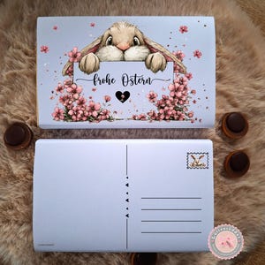 Pode incluir: Um postal de Páscoa azul claro com uma ilustração de um coelho segurando uma placa com a frase "frohe Ostern". O postal é decorado com flores cor-de-rosa e um coração. Um selo de coelho está na parte de trás.