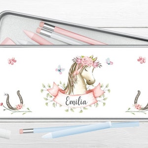 Könnte beinhalten: Eine weiße Bleistiftbox aus Metall mit einer rosa Pferdeillustration, Blumen und Schmetterlingen. Der Name "Emilia" ist auf einem rosa Band geschrieben. Die Box ist geöffnet und enthält Bleistifte.