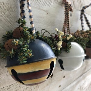 Op de afbeelding: Drie decoratieve metalen belletjes met verschillende kleuren verf en versieringen. Elke bel is versierd met een ander type groen en een lint.