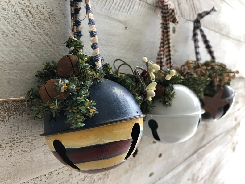 Op de afbeelding: Drie decoratieve metalen belletjes met verschillende kleuren verf en versieringen. Elke bel is versierd met een ander type groen en een lint.