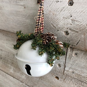 Op de afbeelding: Witte keramische bel ornament met een groene en bruine dennentak en dennenappels eraan bevestigd. De ornament hangt aan een rood-wit geruit lint.
