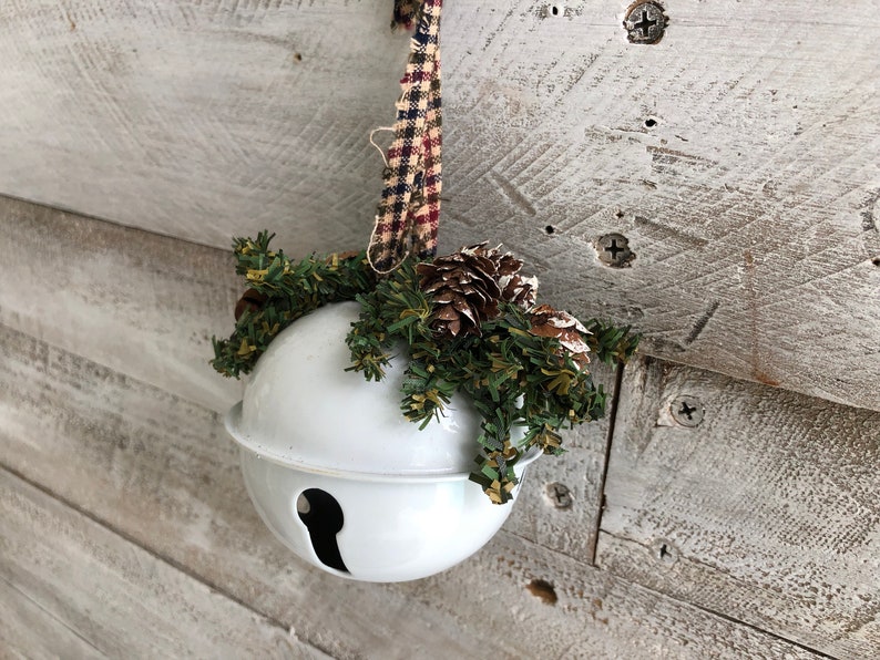 Op de afbeelding: Witte keramische bel ornament met een groene en bruine dennentak en dennenappels eraan bevestigd. De ornament hangt aan een rood-wit geruit lint.