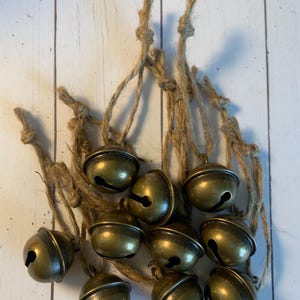 May include: A collection of small, antique brass bells hanging from natural twine. The bells have a classic, rounded shape with a dark, aged finish. The twine is knotted at the top, suggesting they are ready to hang. The background is a light-colored, wooden surface.